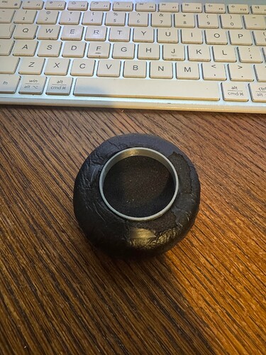 A small, dark-colored, leather-like object with a gray ring in the center sits on a wooden desk in front of a keyboard. (Captioned by AI)