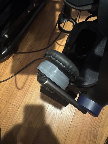 A gray 3D-printed adapter is attached to a microphone and headphones, mounted on a dark-colored device resting on a wooden floor. (Captioned by AI)